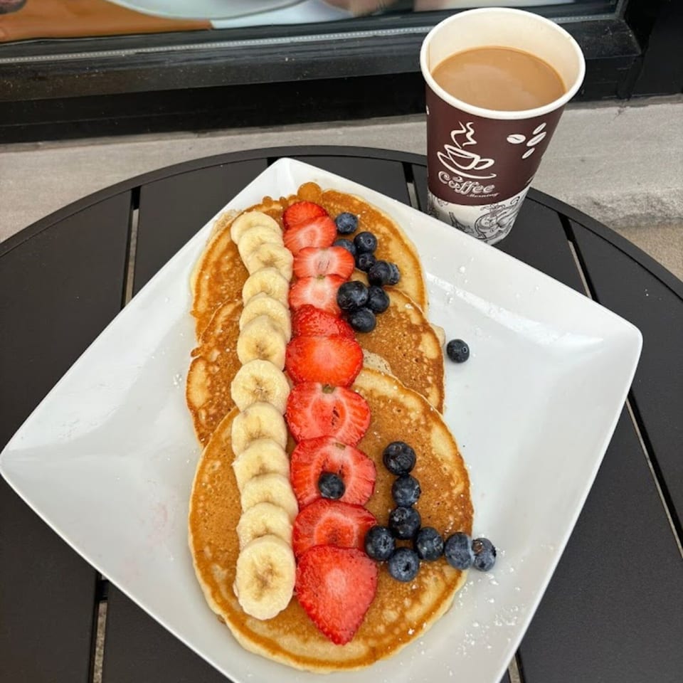 Pancakes topped with sliced bananas, strawberries, and blueberries, served with a cup of coffee.