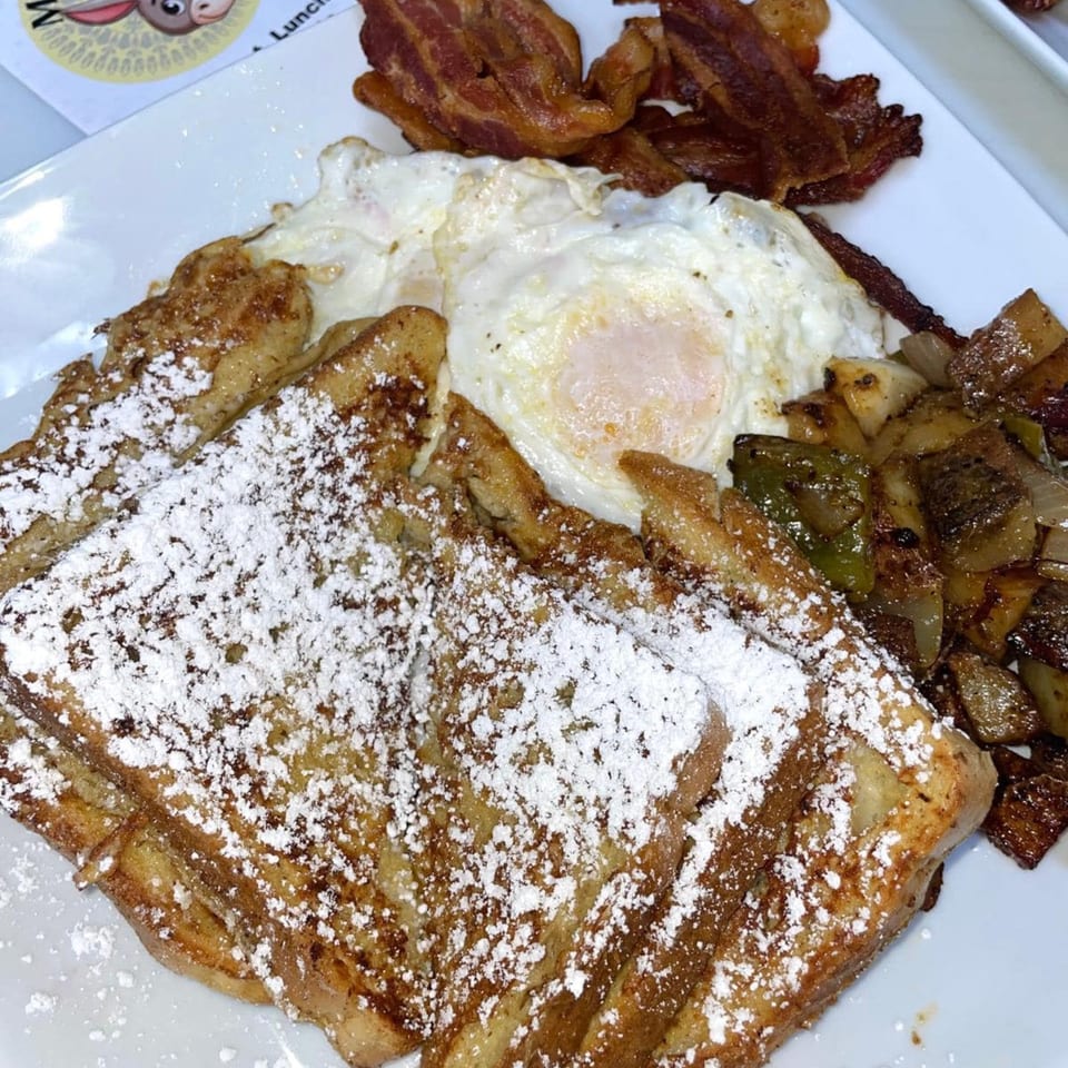 A breakfast plate with powdered sugar French toast, two fried eggs, bacon, and roasted potatoes.