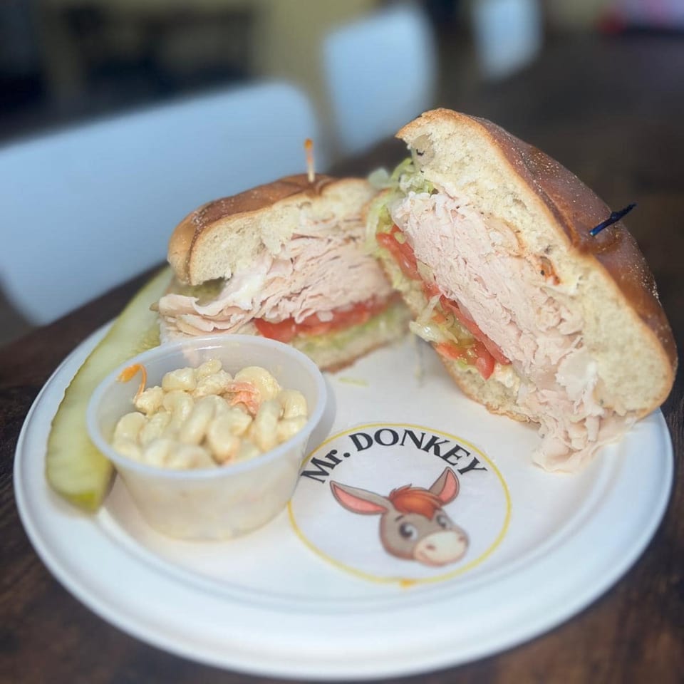 A Mr. Donkey turkey sandwich with lettuce and tomato, served with macaroni salad and a pickle.